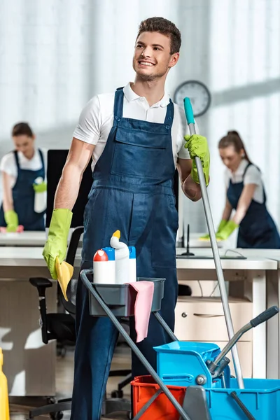 Limpador sorridente em pé perto do carrinho com suprimentos de limpeza e olhando para longe — Fotografia de Stock