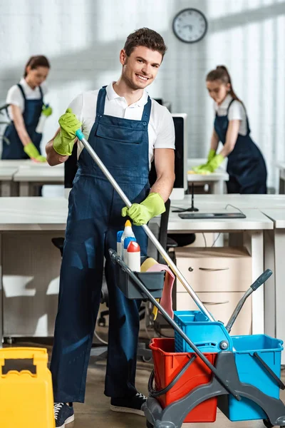 Nettoyant gai debout près du chariot avec des fournitures de nettoyage et souriant à la caméra — Photo de stock