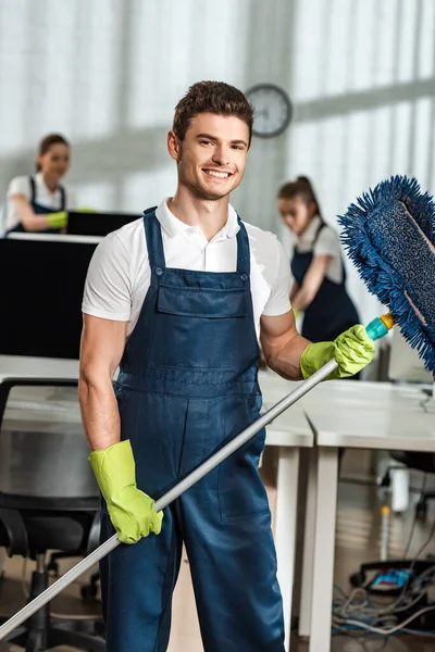 Guapo, limpiador sonriente sosteniendo fregona mientras mira a la cámara - foto de stock