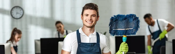 Panoramic shot of handsome cleaner holding mop while smiling at camera near multicultural colleagues — Stock Photo