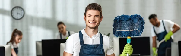 Fotografía panorámica de limpiador guapo sosteniendo fregona mientras sonríe a la cámara cerca de colegas multiculturales - foto de stock