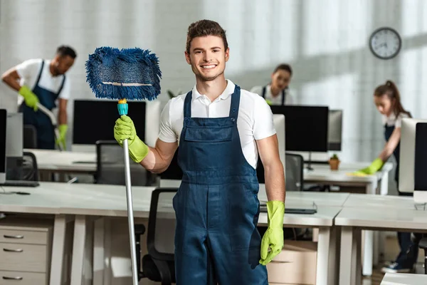 Fröhliche Putzfrau hält Wischmopp in die Kamera neben multikulturellen Kollegen — Stockfoto