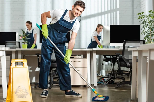 Limpiador joven mirando a la cámara mientras lava el piso con fregona en la oficina moderna - foto de stock