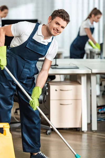 Lächelnde Putzfrau blickt beim Fußbodenwaschen im Büro in die Kamera — Stockfoto