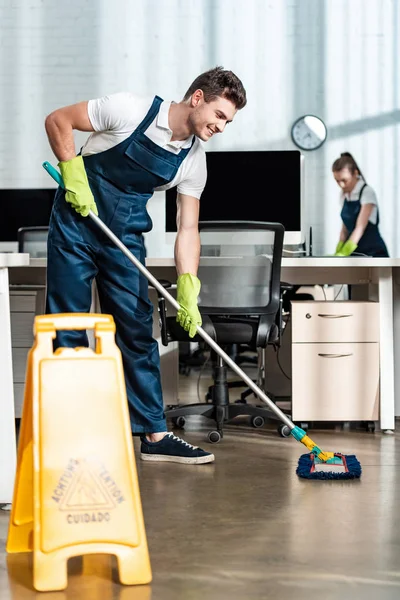 Lächelnd sauberer Waschboden mit Wischmopp in der Nähe des Kollegen Putztisch — Stockfoto