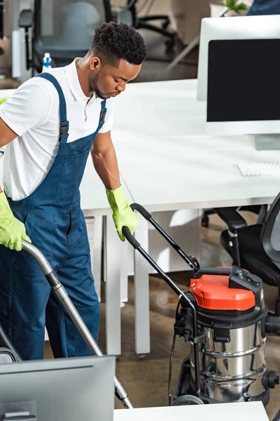 Jeune homme afro-américain nettoyage plancher dans le bureau avec aspirateur — Photo de stock