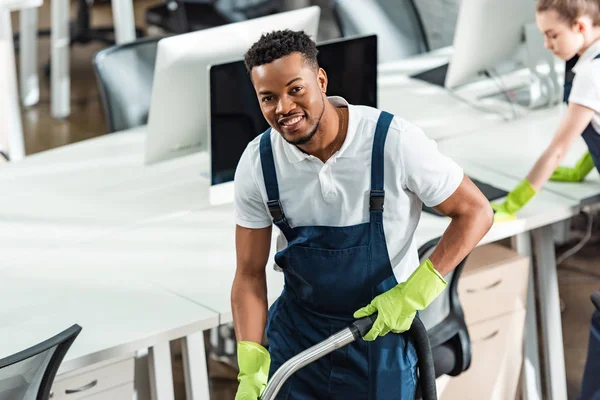 Smiling african american cleaner looking at camera near young colleague — Stock Photo