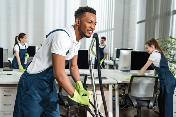 Sonriente africano americano limpiador movimiento aspiradora cerca de equipo de colegas - foto de stock