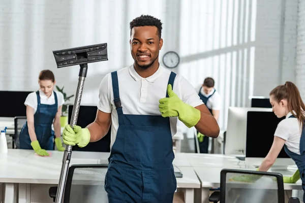 Smiling african american cleaner holding vacuum cleaner brush and showing thumb up near team of colleagues — Stock Photo