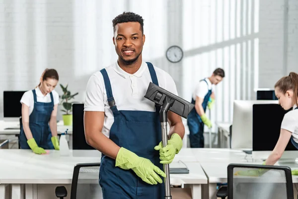 Sorrindo afro-americano limpador segurando escova aspirador perto da equipe de colegas multiculturais — Fotografia de Stock