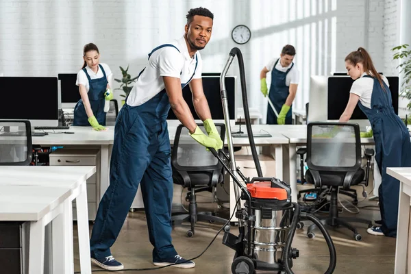 Lächelnde afrikanisch-amerikanische Putzfrau bewegt Staubsauger neben Team multikultureller Kollegen — Stockfoto