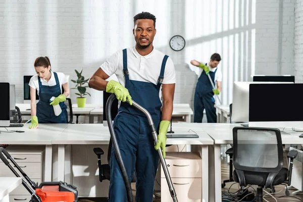 Gutaussehende afrikanisch-amerikanische Putzfrau schaut in die Kamera, während sie mit Staubsauger in der Nähe von Kollegen steht — Stockfoto