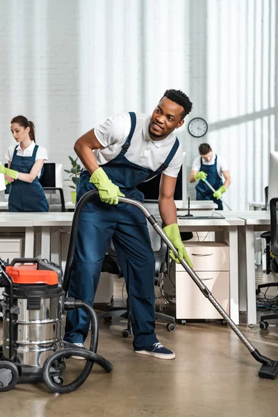 Sonriente africano americano limpiador aspirar piso mientras mira hacia otro lado - foto de stock