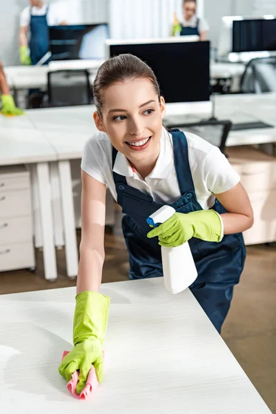 Limpador sorridente segurando frasco de spray enquanto lavava a mesa com pano e olhando para longe — Fotografia de Stock