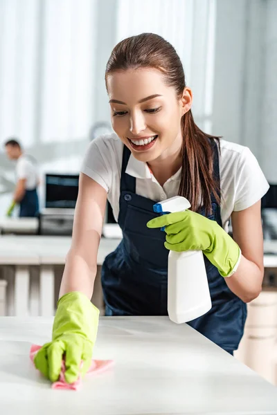 Счастливая уборщица держит в руках баллончик со спреем во время мытья стола тряпкой — стоковое фото