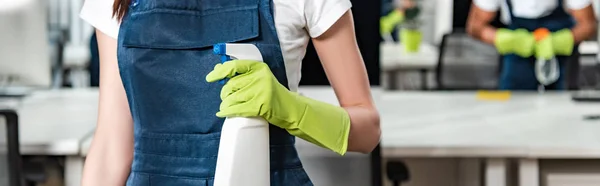 Cropped view of young cleaner in overalls holding spray bottle with detergent — Stock Photo