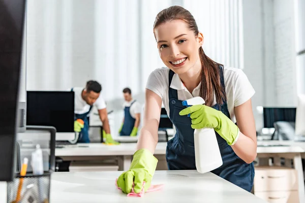 Selective focus of smiling cleaner washing office desk with rag — Stock Photo