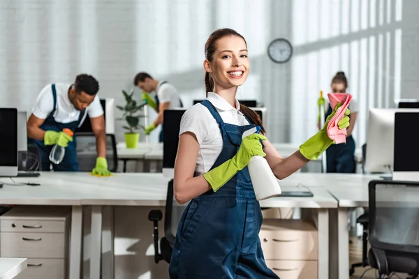 Happy cleaner holding detergent spray and rag while standing near multicultural colleagues and looking at camera — Stock Photo
