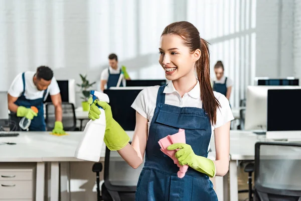 Happy, attractive cleaner holding detergent spray and rag while standing near multicultural colleagues — Stock Photo