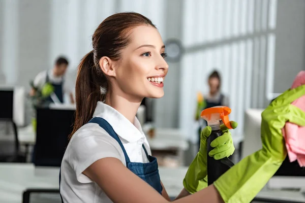 Limpiador feliz y atractivo que sostiene el detergente en aerosol y trapo mientras limpia la oficina - foto de stock