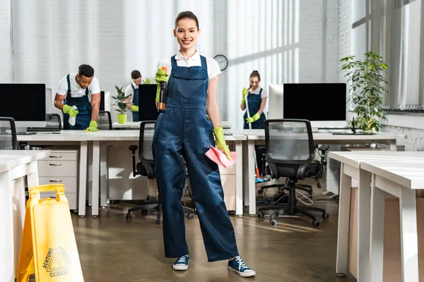 Smiling cleaner holding spray and rag while standing near working multicultural colleagues — Stock Photo