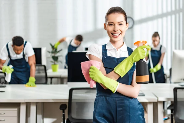 Nettoyeur heureux avec spray et chiffon souriant à la caméra tout en se tenant près de collègues multiculturels — Photo de stock