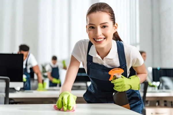 Glückliche Putzfrau schaut in die Kamera, während sie Büroschreibtisch mit Lappen säubert — Stockfoto