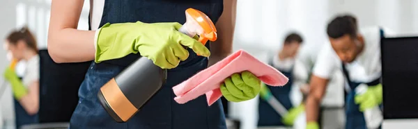 Cropped view of cleaner in uniform spraying detergent on rag, panoramic shot — Stock Photo