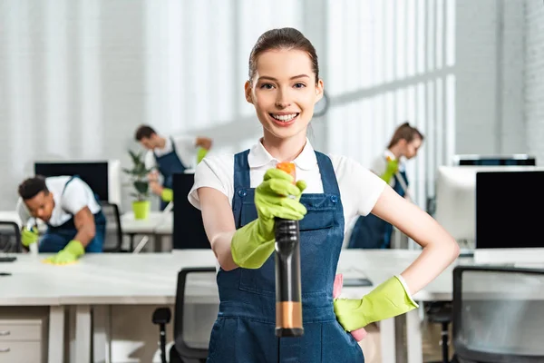 Cheerful cleaner in overalls standing with hand on hip while holding spray bottle and looking at camera — Stock Photo