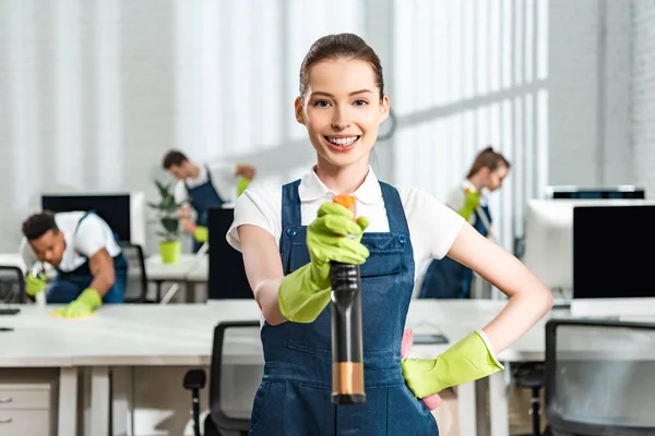 Fröhliche Putzfrau in Overalls, die mit der Hand auf der Hüfte steht, während sie die Sprühflasche hält und in die Kamera schaut — Stockfoto