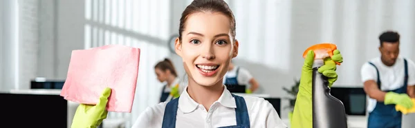 Panoramic shot of happy cleaner holding spray bottle and rag while looking at camera — Stock Photo