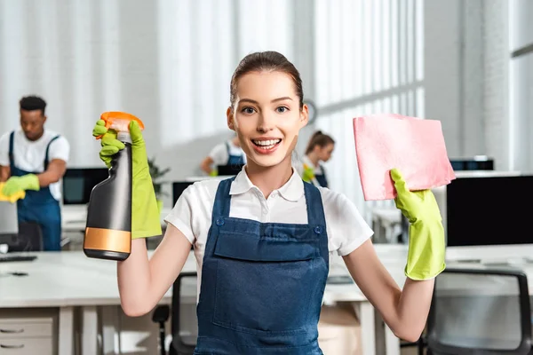 Limpiador feliz en overoles sosteniendo botella de spray y trapo mientras mira a la cámara — Stock Photo