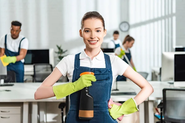 Glückliche Reinigungskraft, die mit der Hand auf der Hüfte steht und die Sprühflasche hält, während sie in die Kamera schaut — Stockfoto