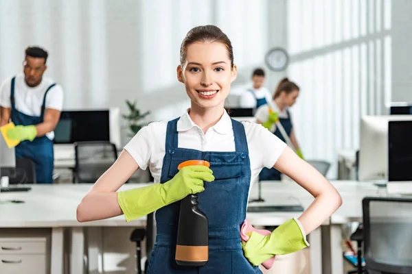 Limpiador feliz de pie con la mano en la cadera y la celebración de botella de spray mientras mira a la cámara - foto de stock