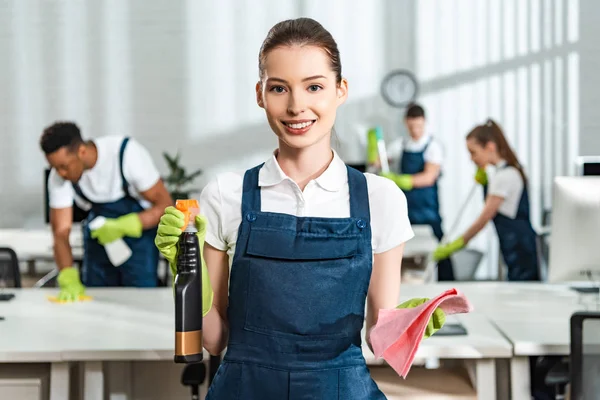 Attractive, happy cleaner holding spray bottle and rag while looking at camera — Stock Photo