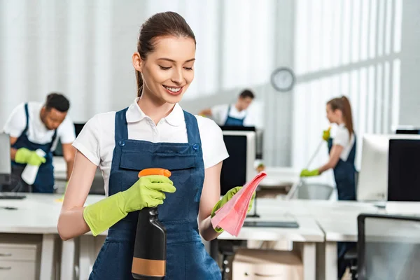 Attraktiver, lächelnder Reiniger, der Waschmittel auf Lappen sprüht — Stockfoto