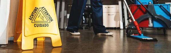 Cropped view of cleaner washing floor with mop near wet floor caution sign, panoramic shot — Stock Photo