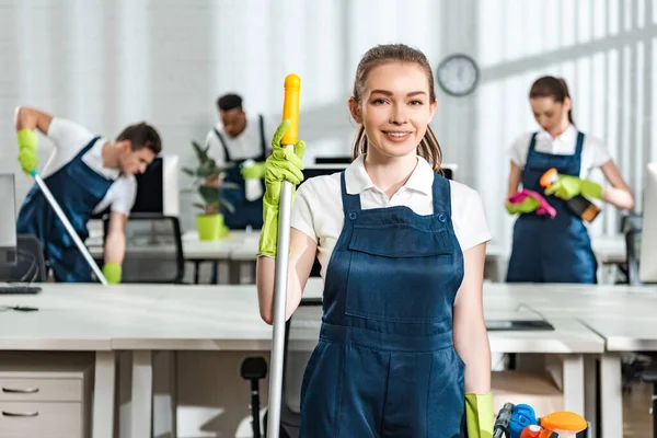Smiling cleaner looking at camera while standing near team of multicultural colleagues — Stock Photo