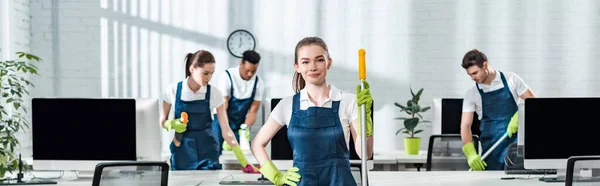 Plano panorámico de limpiador sonriente de pie con la mano en la cadera cerca de colegas multiculturales - foto de stock