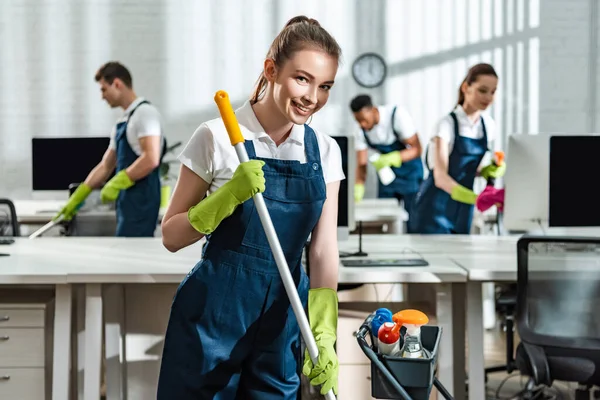Smiling cleaner washing floor and smiling at camera near multicultural colleagues — Stock Photo