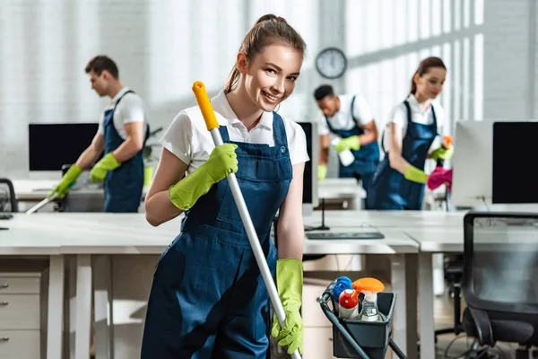 Limpiador sonriente lavando el piso y sonriendo a la cámara cerca de colegas multiculturales - foto de stock