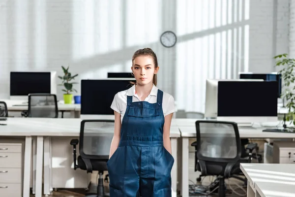 Ernsthafte junge Putzfrau in Overalls blickt in die Kamera, während sie die Hände in den Taschen hält — Stockfoto