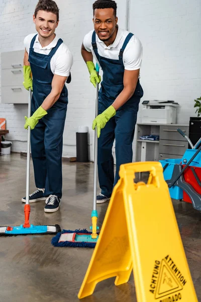 Limpiadores multiculturales sonrientes lavando el suelo con fregonas cerca del suelo húmedo signo de precaución - foto de stock