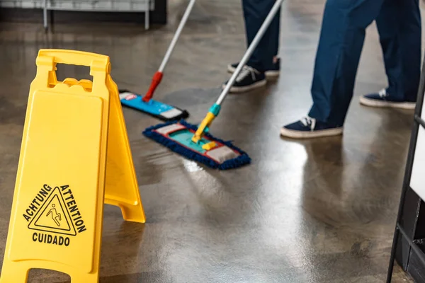 Partial view of two cleaners washing floor with mops near wet floor caution sign — Stock Photo