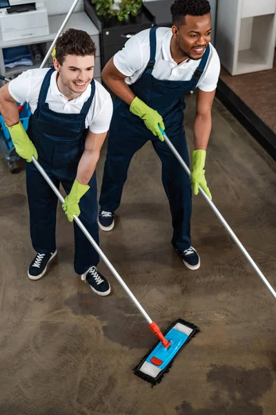 Dos limpiadores multiculturales sonrientes lavando el piso con fregonas - foto de stock