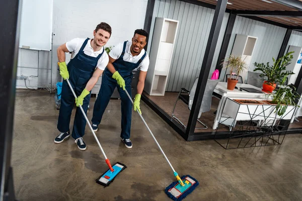 Dos limpiadores multiculturales sonrientes lavando el piso con trapeadores y mirando a la cámara - foto de stock