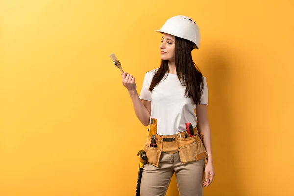 Sério handywoman segurando e olhando para pincel de tinta no fundo amarelo — Fotografia de Stock