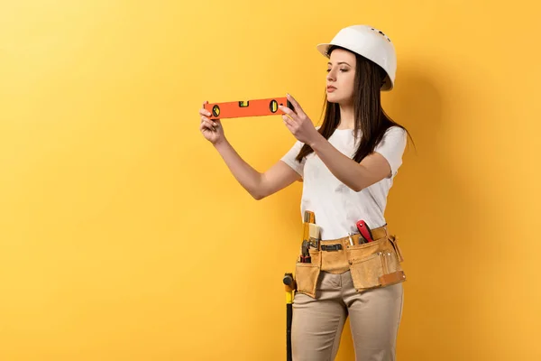 Manitas concentradas en casco sosteniendo el nivel de espíritu sobre fondo amarillo - foto de stock