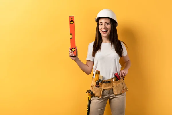 Lächelnde Handarbeiterin im Helm mit Wasserwaage auf gelbem Hintergrund — Stockfoto