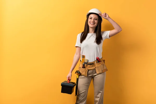 Sonriente manitas en casco sosteniendo caja de herramientas sobre fondo amarillo - foto de stock